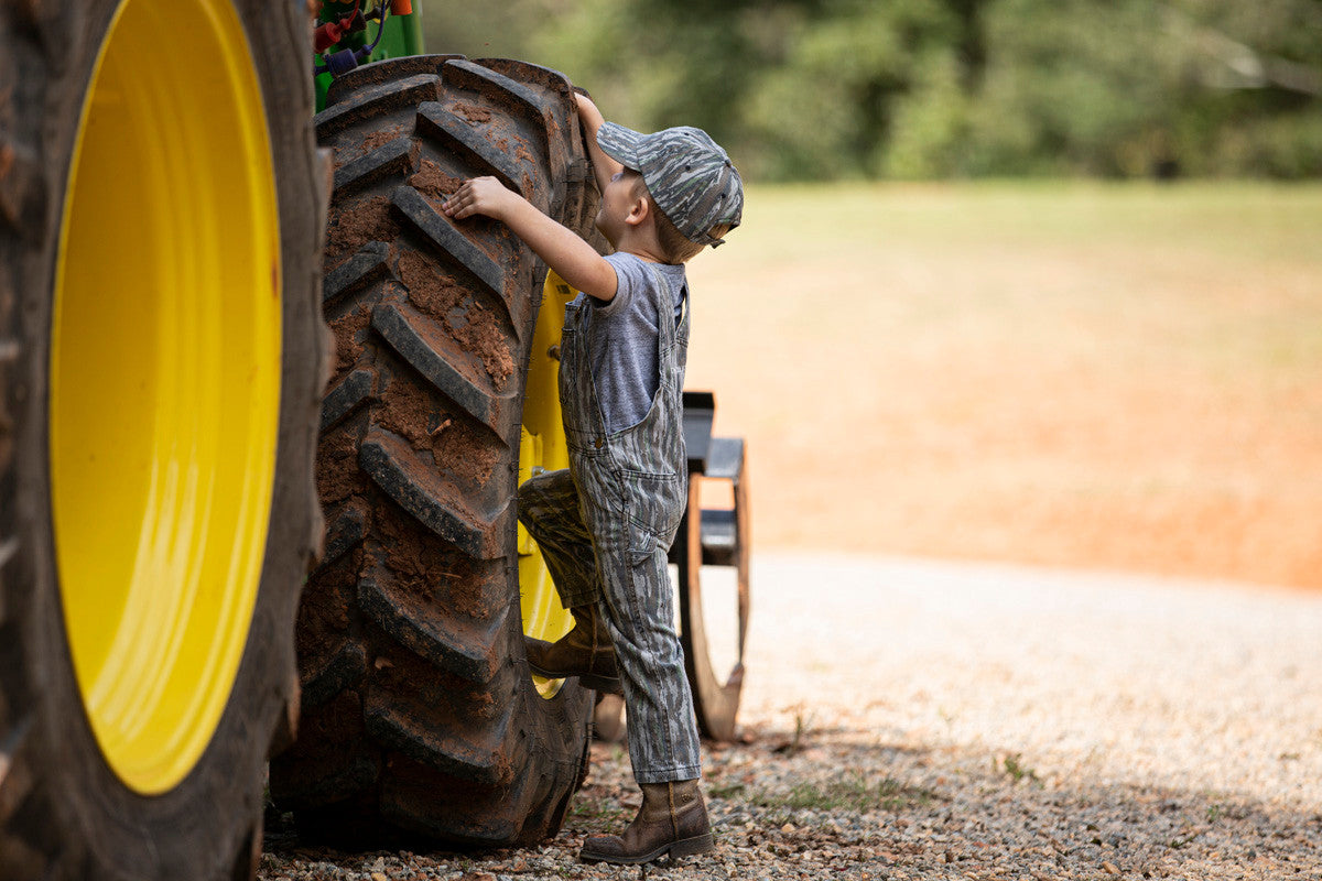 toddler climbing tractor in bc raskulls camo twill overalls and camo cap